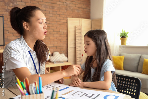 Female Asian speech therapist with logopedic probe and little girl at table i...