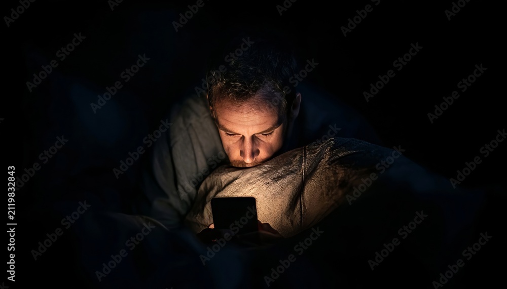 Obraz premium Man Browsing on Smartphone Late at Night in Cozy Bedroom, Engaging with Social Media and News