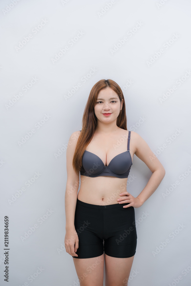 Fototapeta premium Confident Woman Posing in Undergarments: A young woman with long, brown hair stands confidently, wearing a grey bra and black shorts against a light grey background.