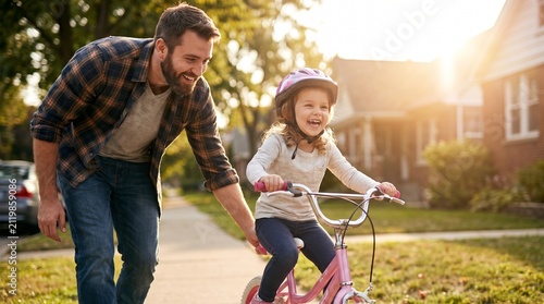 Happy father teaching his young daughter to ride a bicycle on a sunny suburban sidewalk during golden hour