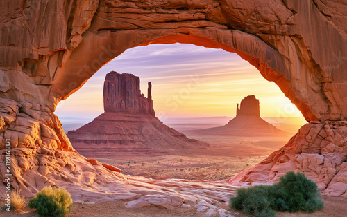 Golden sunrise light illuminates red sandstone rock arch in desert canyon. Massive monolith formation stands in distance during dawn.