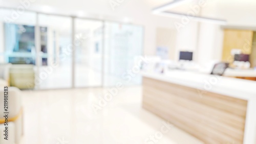 Blur abstract background of corridor in clean hospital. Blurry view of aisle in office with light floor. Defocus walkway in white light building hall with patient registration at reception desk.