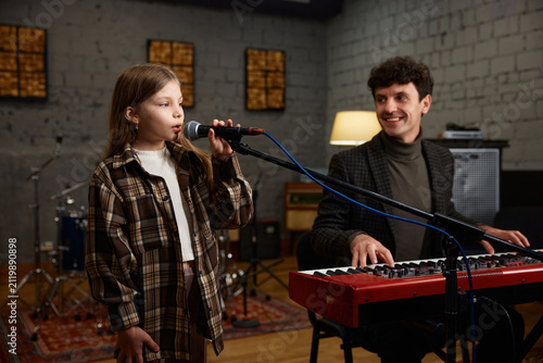 Coach accompanies girl vocalist at mic during focused studio rehearsal