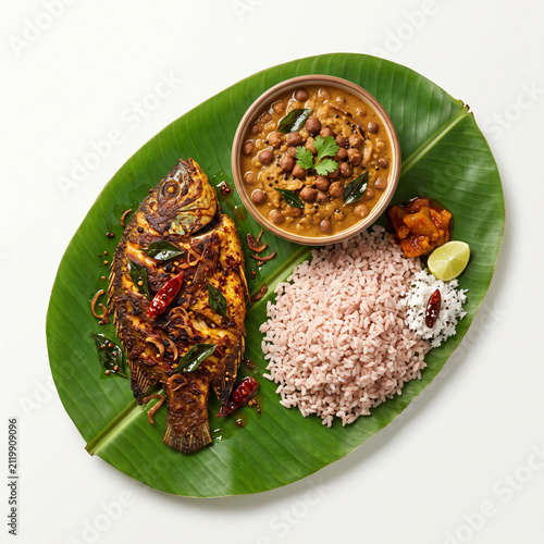 A traditional South Indian meal featuring spicy fish curry, red rice, and chickpeas served on a natural green banana leaf.