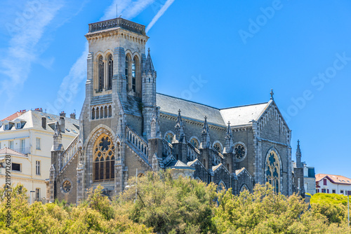 Low-angle view looking up at the historic stone facade and bell tower of Église Sainte-Eugénie under a bright blue sky in Biarritz, France