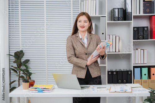 Cuadro en lienzo Ambitious Woman in Office: A young businesswoman with a confident smile stands at her desk in a modern office, surrounded by organized files and a laptop, ready to tackle her tasks