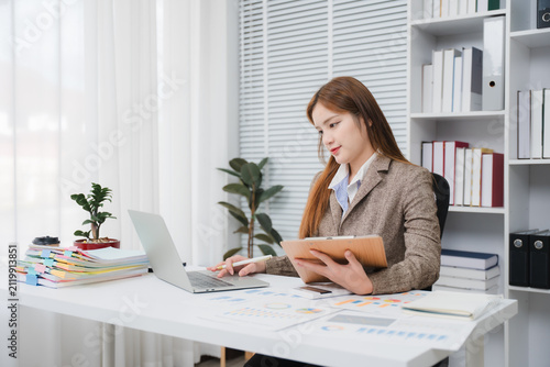 Картината върху платно Focused professional at her desk: A young businesswoman attentively works on her laptop while holding a folder, surrounded by office supplies and neatly organized shelves