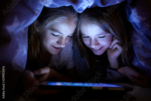 Two sisters playing secretly with their digital tablet, hidden under blanket