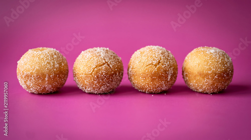 Four round sugarcoated dough balls lined up neatly on a solid pink surface