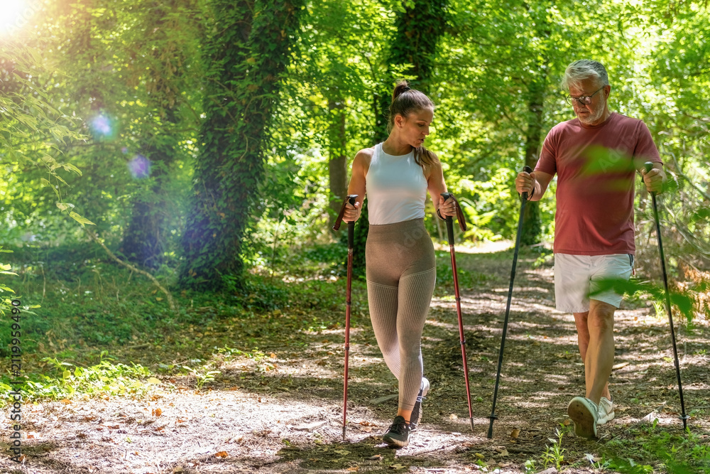 Fototapeta premium Fitness coach leading a senior through a Nordic walking session designed to improve walking mechanics, aerobic capacity, balance, coordination, and long-term physical wellness outdoors.