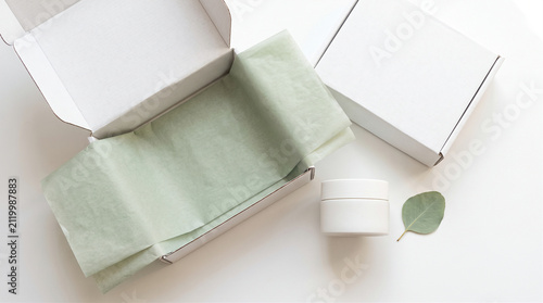Flat lay of blank white cosmetic jar and shipping box with green tissue paper, eco-friendly e-commerce unboxing mockup with copy space