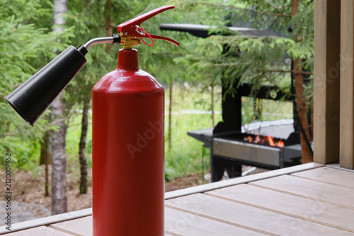 Red fire extinguisher in a wooden house near a barbecue and against the backdrop of a forest. Selective focus