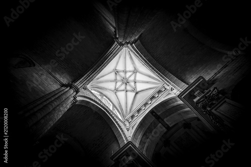 The ceiling of an ancient Gothic cathedral with dramatic lighting, a black-and-white architectural perspective from below. Black-and-white horizontal photo