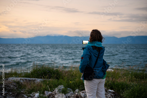 Female tourist taking landscape photograph of lake and mountains with smartphone at sunset