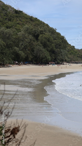 The image depicts a tranquil beach scene alongside a large, densely vegetated hill or small mountain. The sandy shoreline extends into the water, with gentle waves lapping against the shore