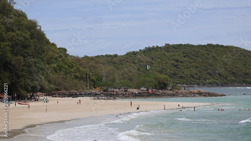 The image depicts a peaceful beach scene with a wide stretch of sandy shore bordered by lush, green, tree covered hills