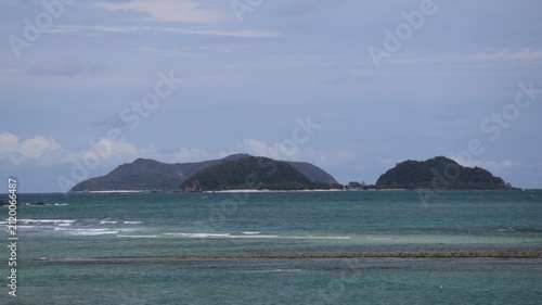 The image depicts a serene seascape with calm, layered waves washing onto the shore. In the distance, a cluster of lush, green islands rises against a cloudy sky, their shapes soft and rounded