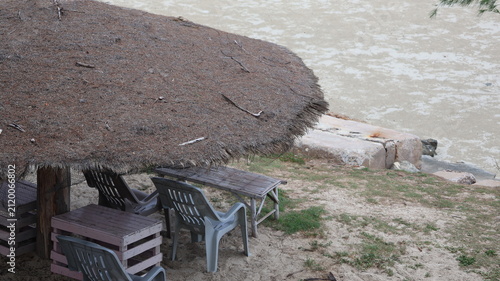 The image depicts a small, outdoor seating area positioned on a sandy surface near a body of water. There are several chairs, including plastic and wooden styles, arranged around a low table