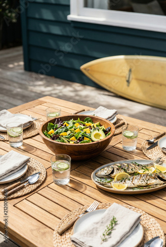 Beautiful summer outdoor dining table set with healthy fresh salad, grilled fish and cold drinks on a wooden deck. A yellow surfboard leans against a blue beach house in the bright sunlight.