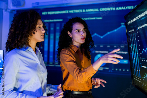 Two adult female business professionals review digital KPI dashboard, discussing annual growth metrics, performance trends, corporate analytics, collaboration, and strategic decision making.