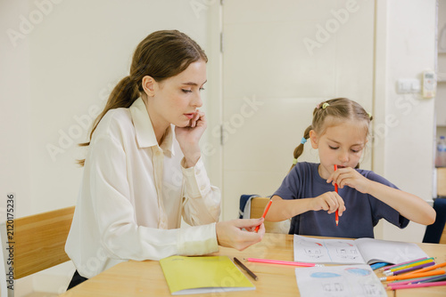 Female teacher guiding a young girl during a colored pencil drawing lesson at a table, representing early education, creative learning, child development, focused study, and classroom support.