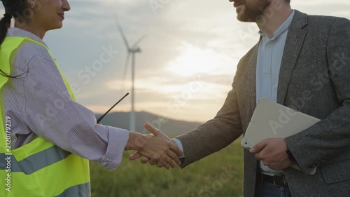 Two professionals shake hands near wind turbines at sunset. Their agreement reflects renewable energy innovation, clean electricity, and sustainable technology driving modern industry and progress.