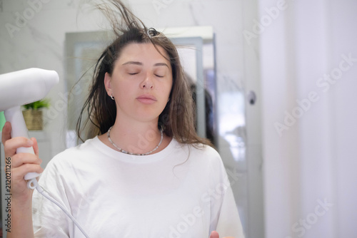 Close-up portrait of a young woman gently blow-drying her hair with a hair dryer, enjoying a relaxing hair care routine with safe drying, healthy styling, and everyday beauty at home.