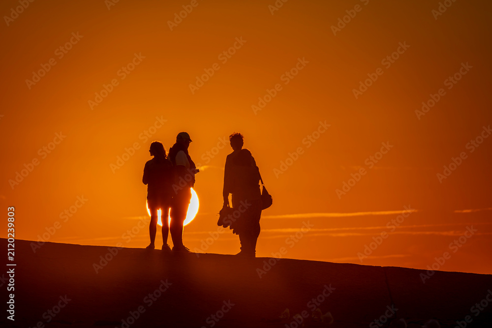 Fototapeta premium Saint-Aubin-Sur-Mer, France - 07 27 2026: View silhouettes of people backlight on the jetty at the beach with the sea and the sun behind at golden hour