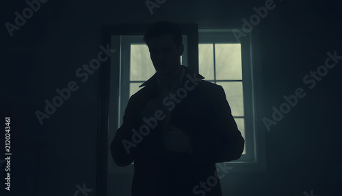 A man is standing in front of a mirror, putting on a jacket. The image has a moody and somewhat mysterious feel to it, as the man is standing in a dimly lit room with a window behind him, realistic.