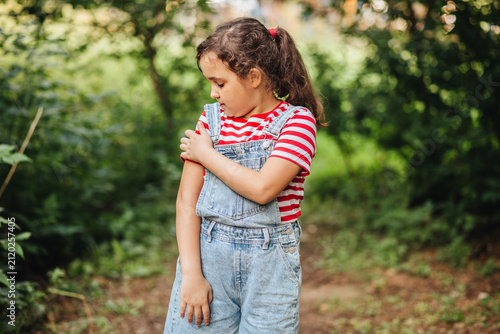 Mosquitoes are biting a girl, and she is scratching her arm in a summer forest. The child, wearing denim overalls and a striped T-shirt, is standing on a forest path.