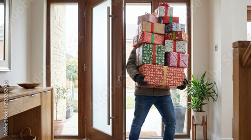 Person carrying an enormous stack of colorful wrapped holiday gifts, struggling to enter or exit a modern home through the front door.