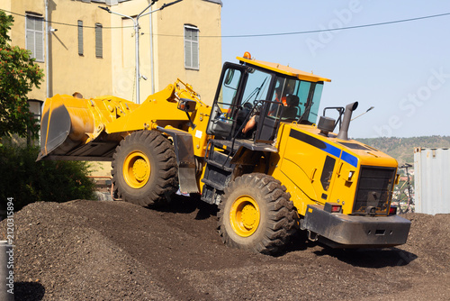 Horizontal street city photo. Yellow bucket loader stands on pile of gravel, sand, pebbles, soil, debris, crushed rock, dirt on construction site. Concept of repair road work, transport, build, job