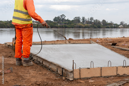 Construction worker applying curing compound with sprayer on fresh concrete slab near riverbank