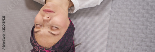 Middle Aged Woman Lying Relaxed With Eyes Closed On Yoga Mat Indoors Close Cropped On Textured Grey Floor Wearing White Shirt And Patterned Scarf Wrapped Around Head During Calm Self Care Moment.