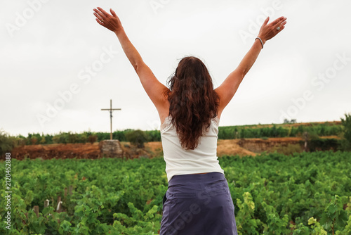 A woman raises her arms toward a cross overlooking vineyards in the Burgundy wine region of France, expressing joy and connection with the peaceful landscape.