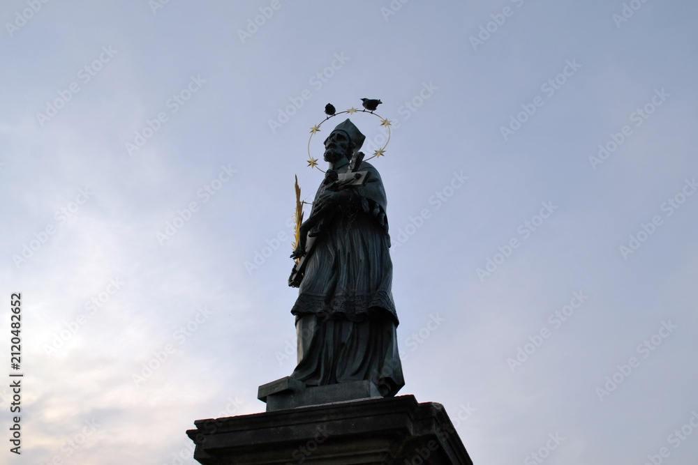 Fototapeta premium Silhouette of a saint statue with halo of stars on a pedestal against dusk sky on charles bridge in Prague for moody background and travel poster design