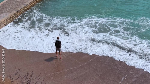 A man wearing a backpack and shorts stands on a sandy beach as turquoise waves roll in and swirl around his ankles. He watches the sea from an elevated coastal viewpoint.
