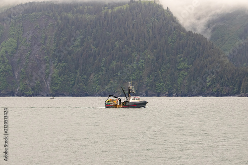 Commercial fishing boat cruising past steep forested mountains on calm ocean water under overcast skies in Kenai Fjords National Park Alaska USA