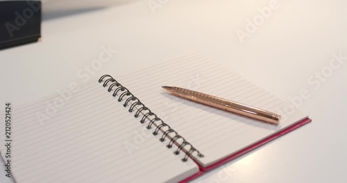 Overhead light is shifting reflections as spiral notebook with rose gold pen is resting on desk