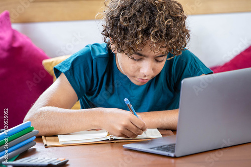 Focused teenage boy studying at home, writing notes in a notebook while using a laptop for online learning. Concentrated student doing homework, preparing for exams or attending a virtual class 