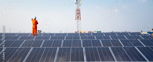 Worker stands tall on solar panels under clear blue skies. Orange suit contrasts with dark panels and distant tower. Sunlight reflects off rows of modern energy collectors