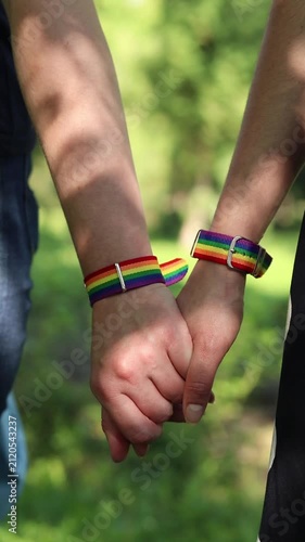 Two people holding hands with LGBTQ+ rainbow bracelets. Close-up of hands holding together with pride symbols. Same-sex couple holding hands on blurred natural background. Love and equality concept