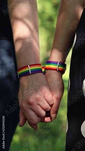 Two people holding hands with LGBTQ+ rainbow bracelets. Close-up of hands holding together with pride symbols. Same-sex couple holding hands on blurred natural background. Love and equality concept