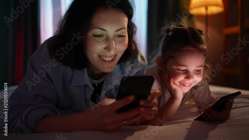 Mother and daughter sitting on sofa using smartphone together in dark living room at evening close up
