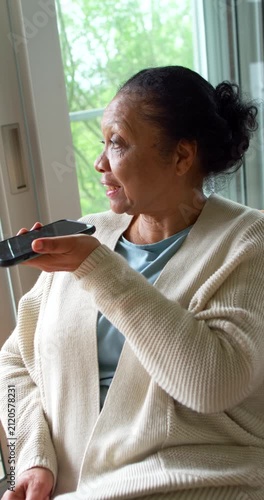 Vertical video: Senior African American woman using smartphone speaker in armchair by sliding door