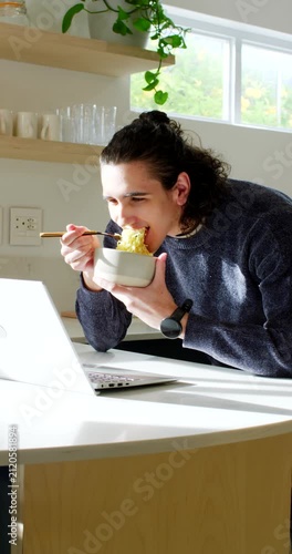 Vertical video: African American man eating noodles with bowl while working on laptop in kitchen