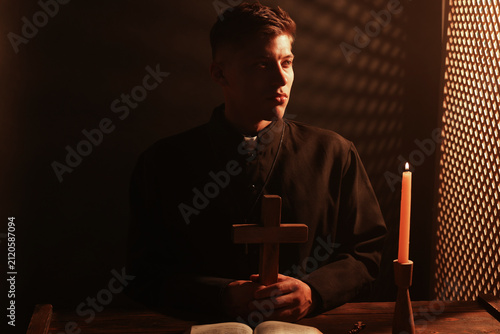 Fotografie Priest holding cross at wooden table with Holy Bible, and candle in confessional
