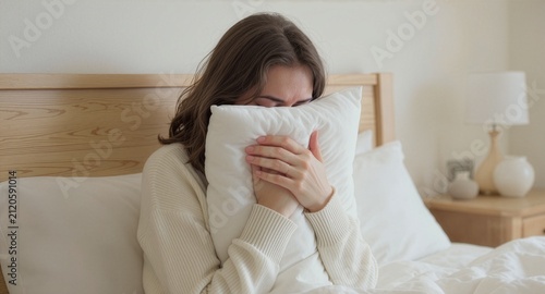 A young woman with brown hair burying her face in a soft white pillow while sitting in bed during a lazy day, embracing a quiet moment of comfort and relaxation inside a cozy bedroom scene