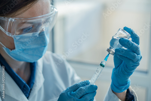Close up of a doctor wearing mask, gloves, and goggles drawing vaccine from vial into syringe. Image highlights modern healthcare, immunization, and infection prevention in a medical environment