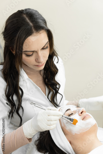 A skilled cosmetologist uses a brush to evenly spread a white facial mask on a woman's face during a spa procedure. The serene atmosphere highlights advanced skincare and wellness trends